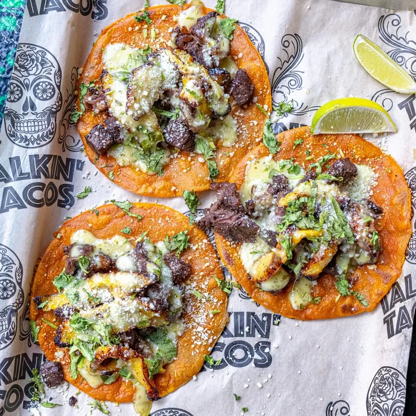Three Talkin' Tacos with meat, cheese, and cilantro on branded paper with lime wedges.