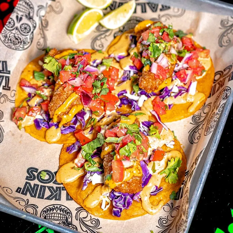 Three Talkin' Tacos with fried fish, purple cabbage, pico de gallo, and sauce, on branded paper.
