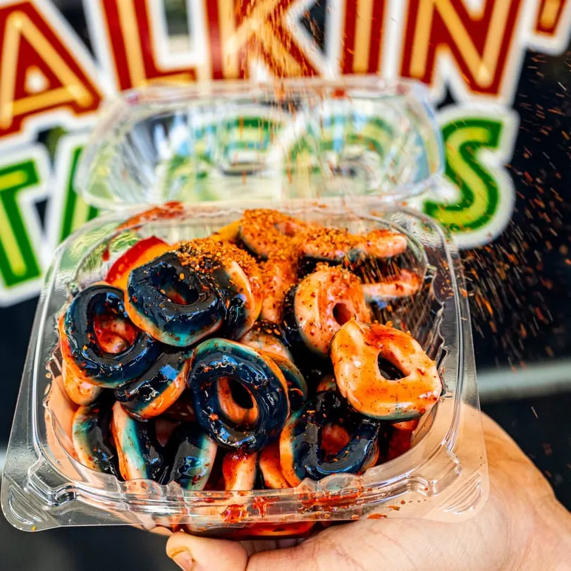 A hand holds a clear plastic container filled with mini donuts, some blue, some orange, covered in chili powder.