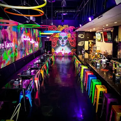Vibrant Talkin' Tacos restaurant interior with colorful chairs, neon lights, and a large Day of the Dead mural.