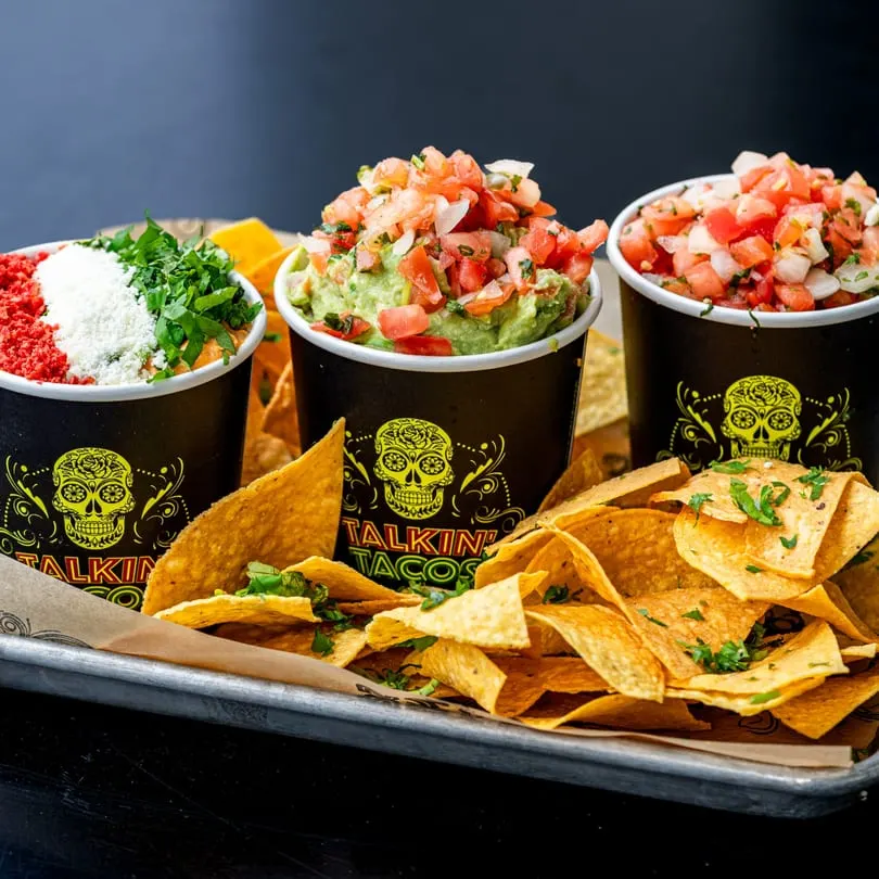 Three Talkin' Tacos cups filled with various toppings and dips, surrounded by tortilla chips on a tray.