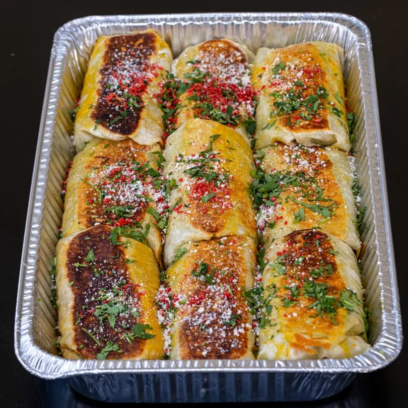 Nine golden-brown burritos, topped with red spice, white cheese, and green herbs, in a foil tray.