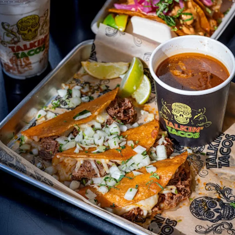 Talkin' Tacos birria tacos with onions, cilantro, lime, and a side of consommé on a metal tray.
