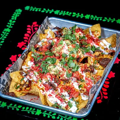 Talkin' Tacos nachos with meat, guacamole, sour cream, and red sprinkles on a black patterned background.