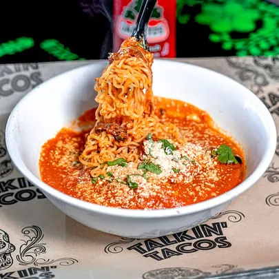 Tongs lifting saucy ramen noodles from a white bowl with red sauce, cheese, and cilantro.