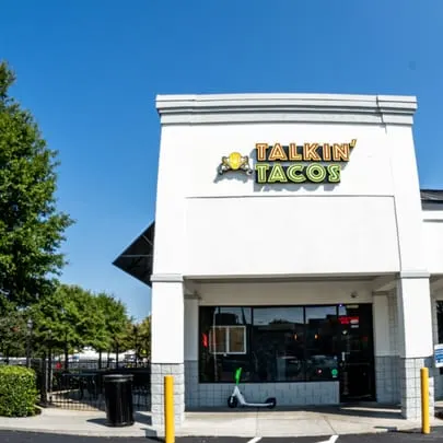 Exterior of Talkin' Tacos restaurant with outdoor seating, a bright sign, and a scooter parked outside.