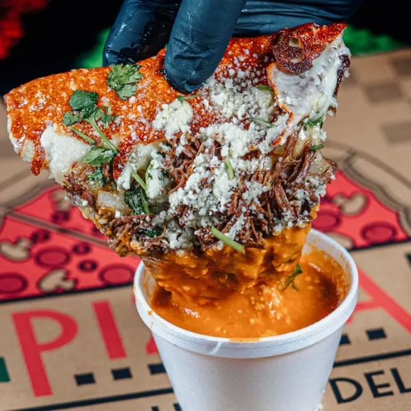 A hand in a black glove dips a cheesy birria quesadilla into a cup of rich consommé.