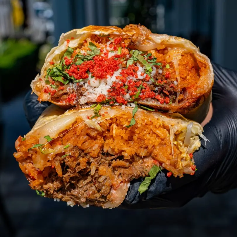 A Talkin' Tacos burrito, cut in half, revealing rice, meat, cheese, and red crunchy toppings.