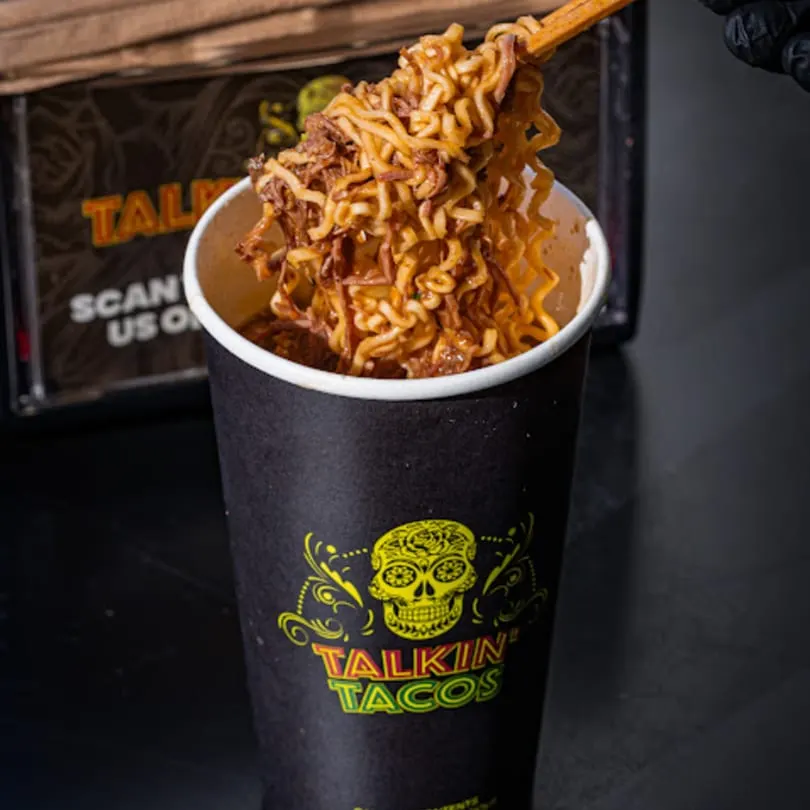 Chopsticks lifting saucy noodles and shredded beef from a black Talkin' Tacos cup.