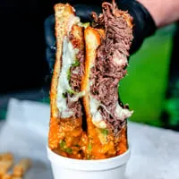 A hand in a black glove holds a large, cheesy birria taco over a cup of consommé.